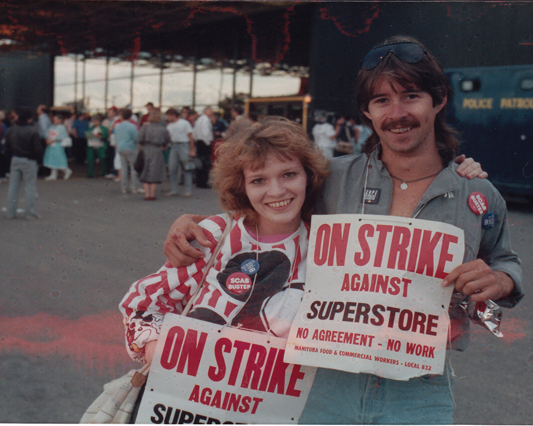 Two people with strike placards pose for photo outside supervalu with striking workers and police paddy wagon in background.