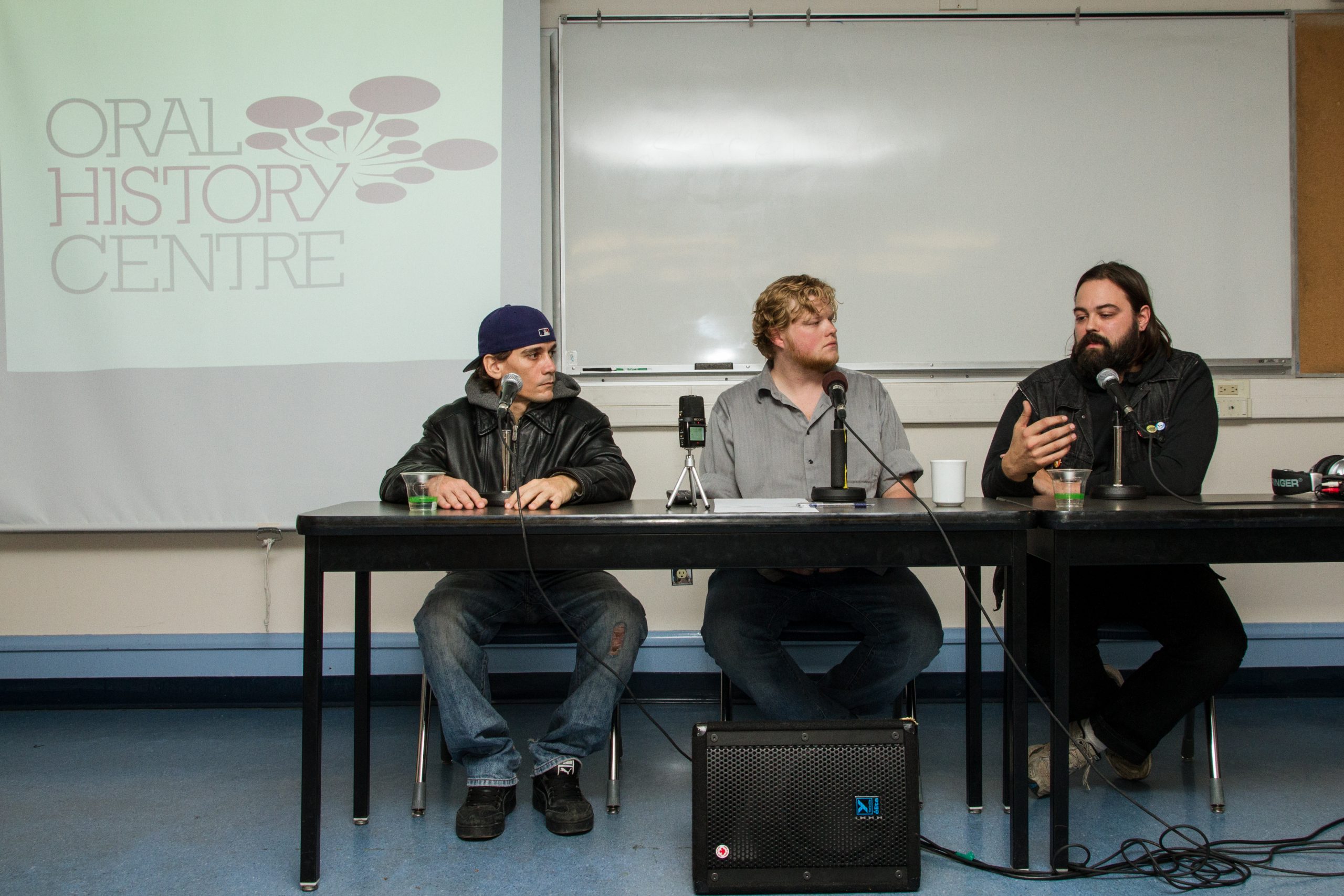 Colour photo of three men at event table with three microphones. Oral History Centre logo is projected behind them.