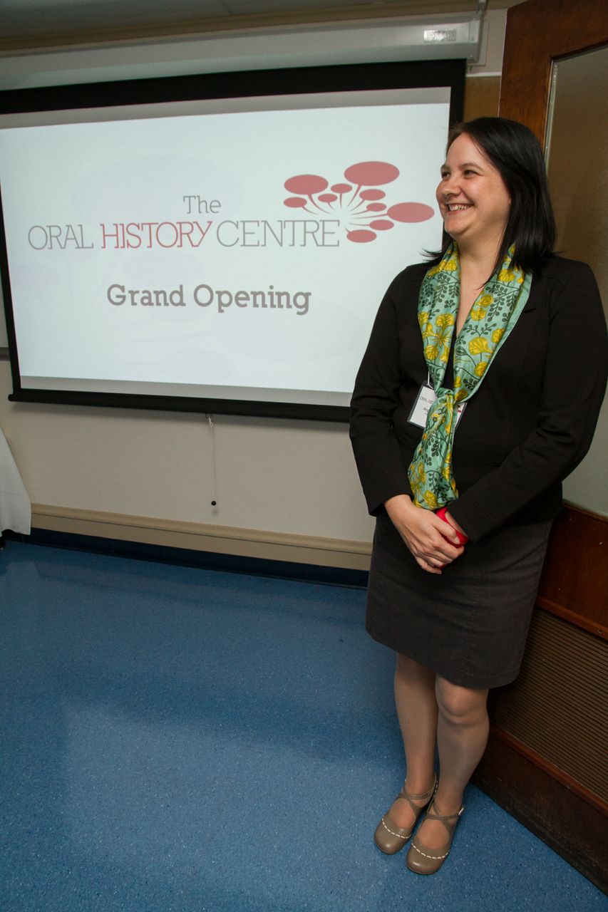 Photo of a woman standing in front of a projection slide with the caption: The Oral History Centre Grand Opening.