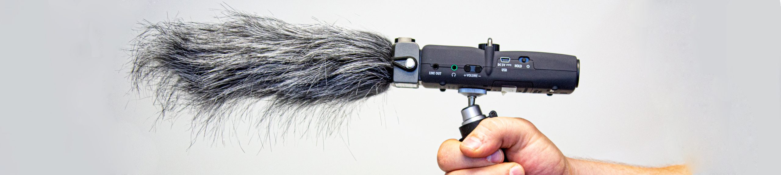 Photo of hand holding grip, digital recorder with shotgun capsule and grey furry wind shield.