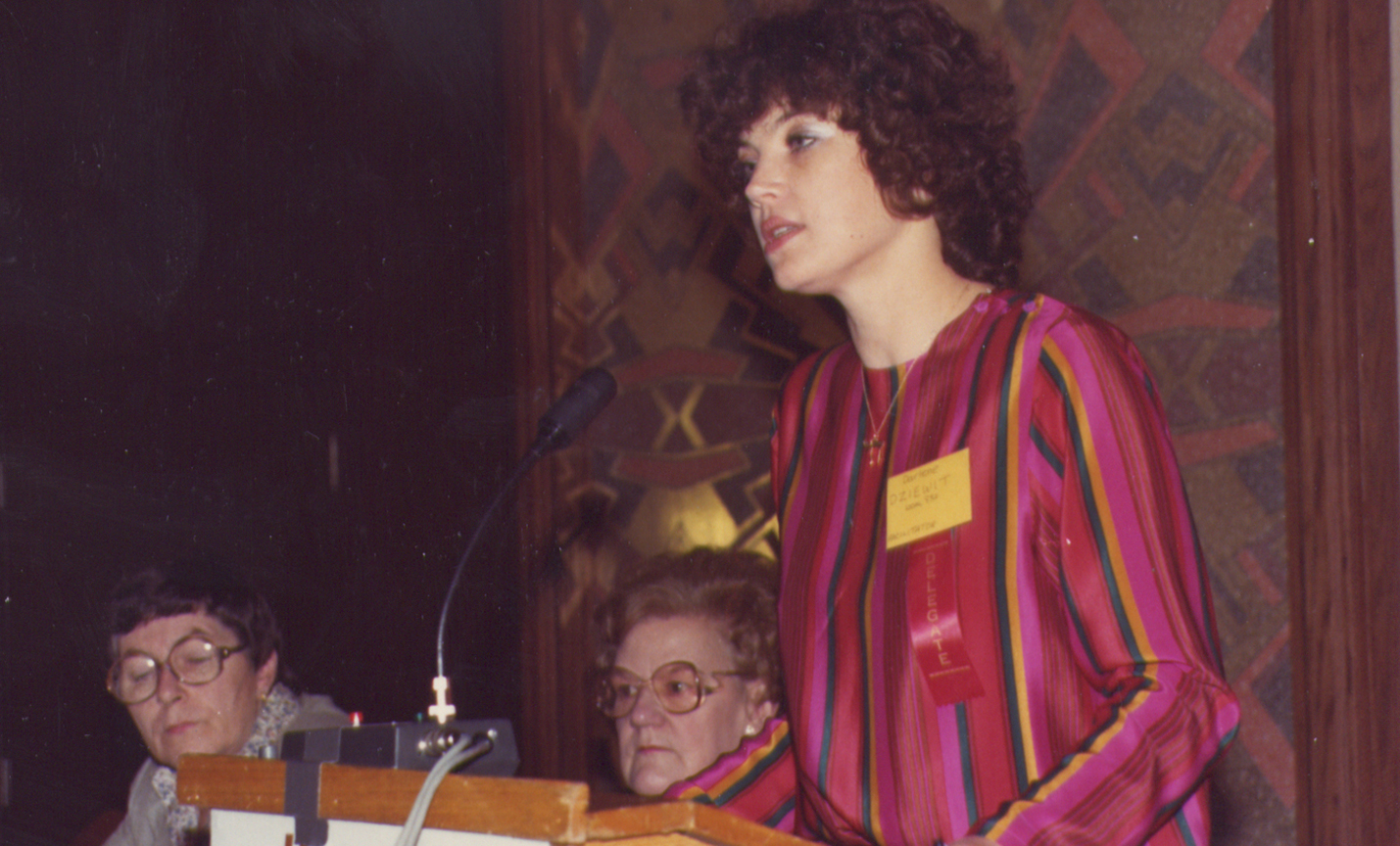 Darlene Dziewit standing at podium, in background next to podium two other women are listening.
