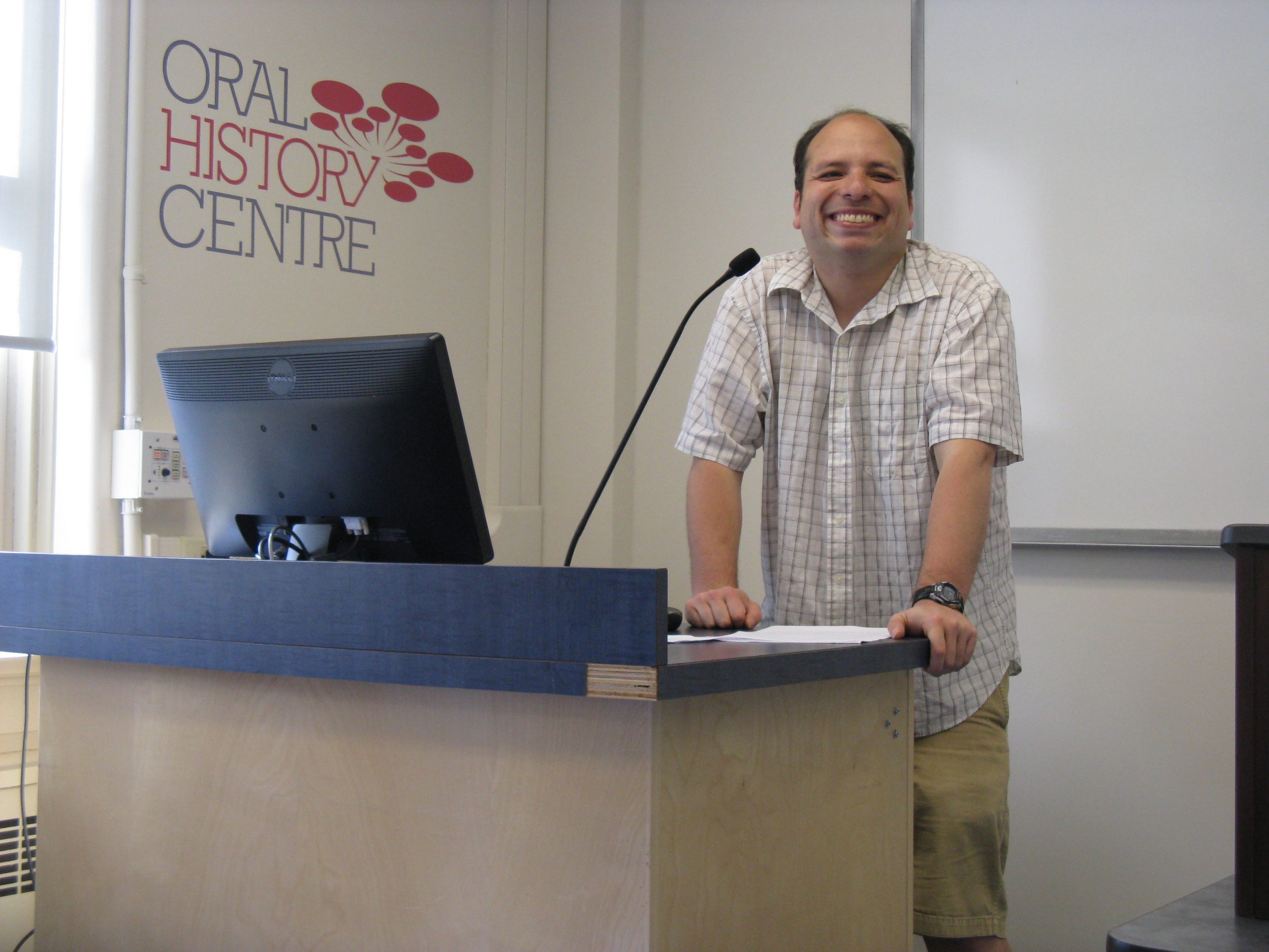 Photograph of Carlos Sosa smiling, standing at podium. In background whiteboard and Oral History Centre logo on wall.