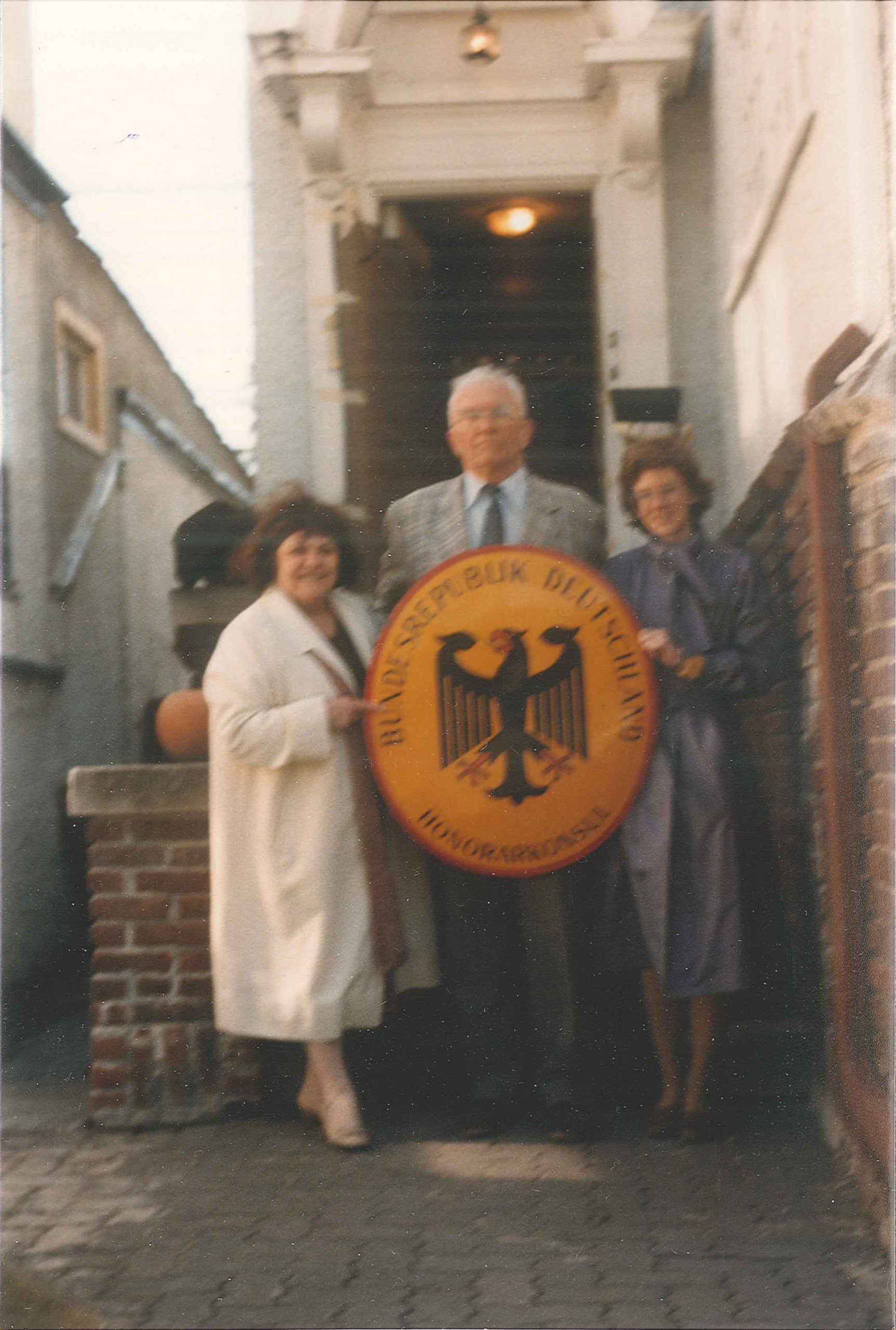 Deborah Spindler, Mrs. Gummelt and Mr. Thiele on the porch of the holding a German coat of arms.