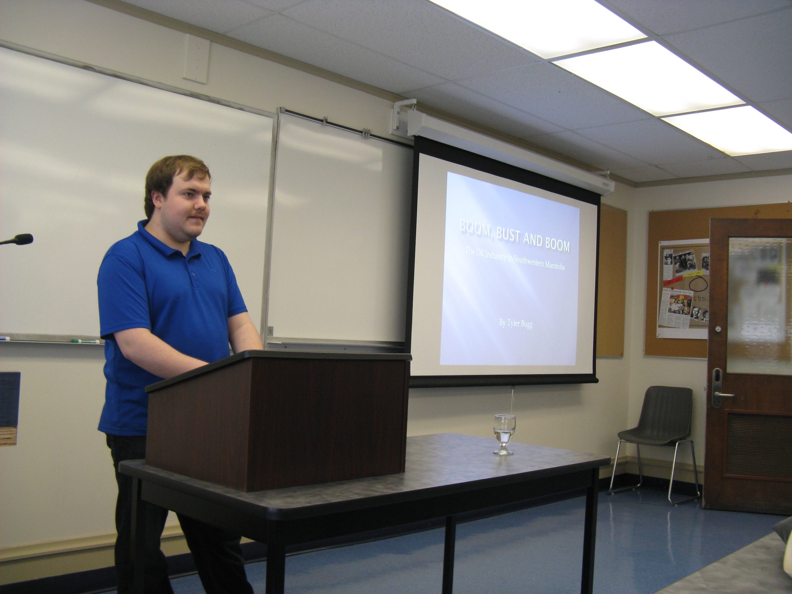 Tyler Bugg stands at small lectern on top of table. Projected powerpoint slide in background. Text: Boom, Bust, and Boom.
