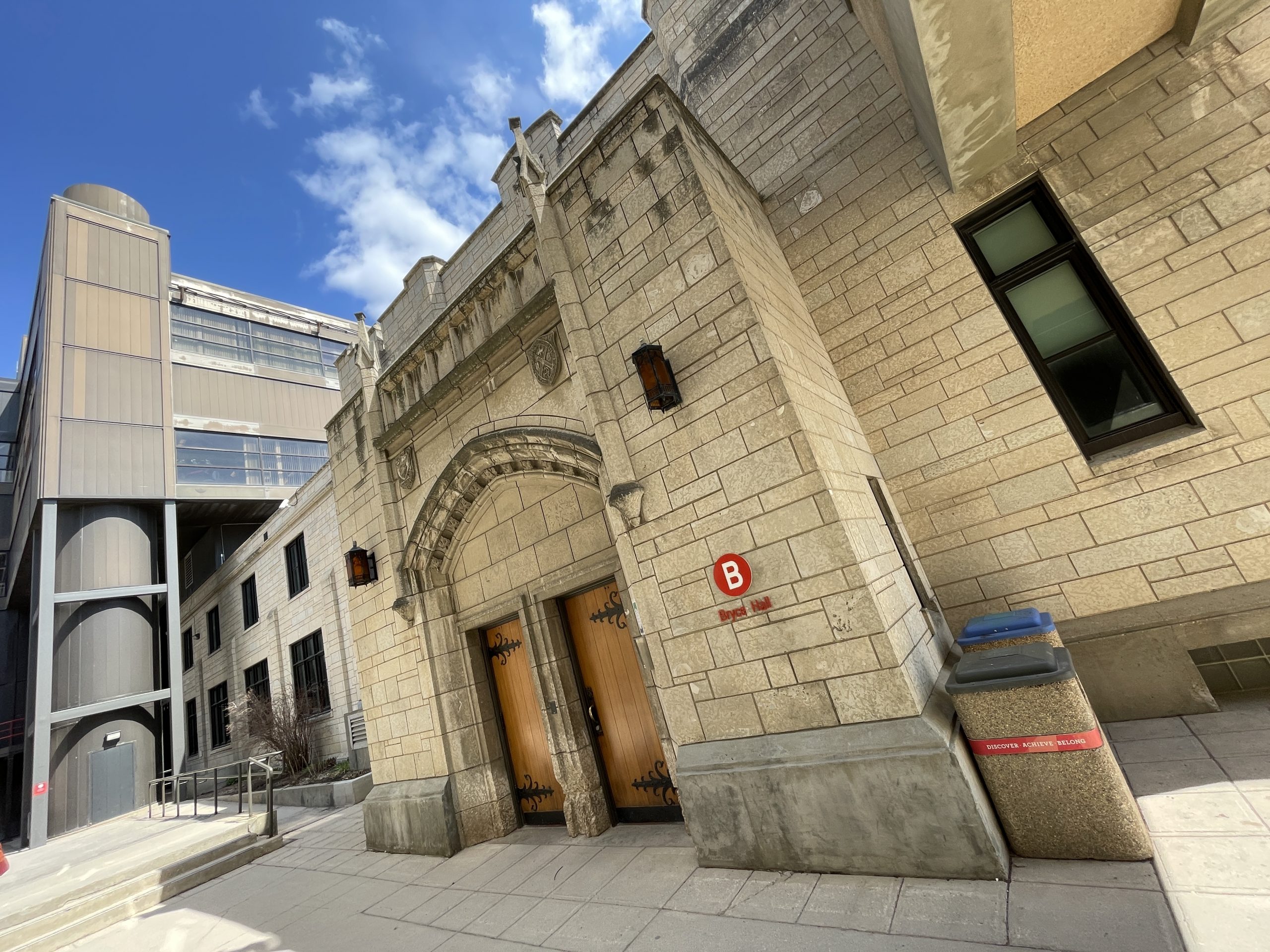 The Tyndall stone exterior and double wooden doors of University of Winnipeg's Bryce Hall in the foreground, with the tubular stairwell of Centennial Hall visible in the background.